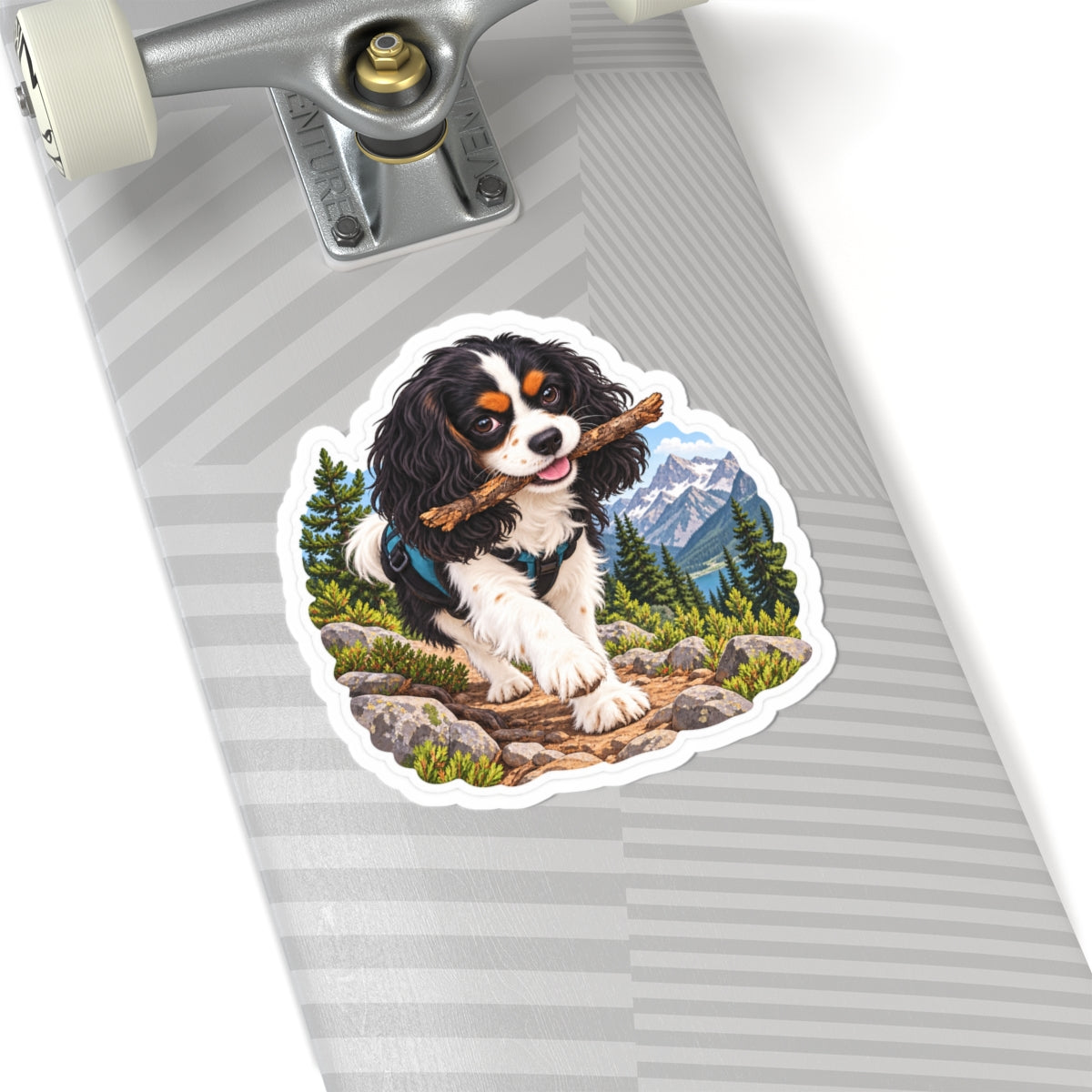 Product mockup of the Cavy Play Mode Trail Stick kiss-cut sticker: a tricolor Cavalier King Charles Spaniel carrying a stick along a mountain trail with pines and snowy peaks.